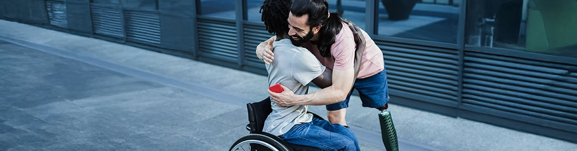 two people hugging, one in a wheel chair and another standing with a prosthetic, because they received Financial Assistance for Prosthetics and Free Prosthetics